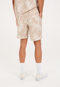 Pantalones cortos beige con estampado de hojas blancas, cinturilla elástica y bolsillos laterales, combinados con calcetines blancos y zapatos claros.