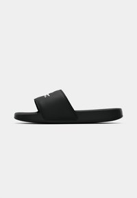 Sandale noire à glissière avec une large bandoulière comportant un petit logo crocodile blanc, semelle plate et design minimal sur fond uni.