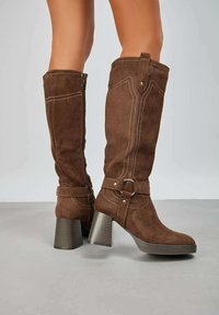 Bottes en suede marron montantes jusqu'au genou, présentant un talon épais et strié, une couture détaillée, et une sangle réglable avec une boucle circulaire.