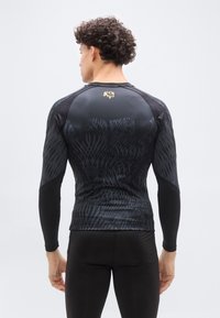 Νέος άν τρας με σγουρά μαλλιά που φοράει μαύρο αθλητικό πουκάμισο με μακριά μανίκια, διακοσμημένο με αφηρημένα σχέδια και ένα μικρό χρυσό εμβλημα στο επάνω μέρος της πλάτης.