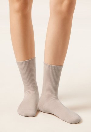 Nohy obuté v světle béžových žebrovaných ponožkách, stojící na světlejším pozadí s mírně rozkročenýma nohama, jednou nohou položenou celou na zemi a druhou s nataženými prsty.