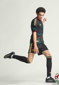 Giovane calciatore maschio in divisa nera con strisce rosse, gialle e verdi mentre calcia un pallone da calcio bianco e rosso su sfondo neutro.
