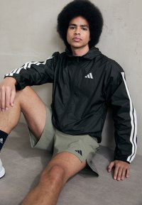 Sort zip-jakke med hvide side-striber og logo, parret med lysegrønne shorts. Mat overflade, ligger på en glat flade.