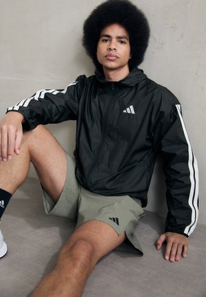 Svart zip-up jakke med hvite sidedetaljer og logo, kombinert med lysegrønne shorts. Matt tekstur, ligger på en jevn overflate.