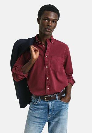 Chemise boutonnée bordeaux avec une poche poitrine, associée à un jean bleu clair et un blazer noir drapé sur une épaule.