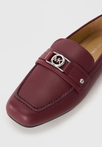 Mocassini in pelle bordeaux con punta quadrata, hardware in metallo argentato con logo MK e dettagli cuciti su una superficie liscia.
