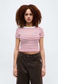 Γυναίκα με κοντά κατσάρα μαλλιά που φοράει λευκό και κόκκινο ριγέ κοντομάνικο crop top και σκούρο καφέ παντελόνι με ψηλή μέση, στέκεται μπροστά σε λευκό φόντο.