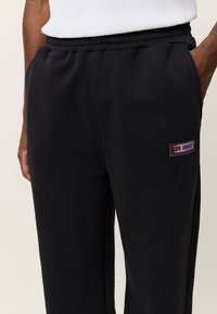 Μαύροι παντελόνες φόρμας από μαλακό ύφασμα, με ρυθμιζόμενη μέση με κορδόνι και μικρό πολύχρωμο λογότυπο στο μηρό.