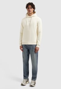 Crèmekleurige sweatshirt met capuchon en een voorkeurszak, gecombineerd met blauwe versleten jeans en lichtgrijze sneakers. Model staat tegen een lichtgrijze achtergrond.