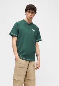 Νεαρός άντρας φόρεμα πράσινο κοντομάνικο πουκάμισο henley και μπεζ παντελόνι cargo, στέκεται με το ένα χέρι στην τσέπη και έχει τατουάζ στο αντιβράχιο.