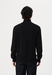 Pull en maille côtelée noir avec un col haut, doté de longues manches et d'un ourlet ajusté, présenté de dos sur un fond uni.