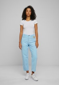 Pantaloni in corduroy blu chiaro con design a gamba dritta, dotati di tasche anteriori e logo ricamato sulla vita. Abbinati a una camicia bianca.