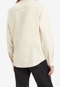 Lichtbeige langemouwen shirt met een puntige schouderpas, knoopmanchetten en een gladde textuur. De achterkant heeft een ronde zoom en subtiele stikdetails.