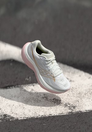 Scarpa da corsa bianca e rosa con tomaia in mesh e logo, sospesa sopra l'asfalto con strisce bianche della strada e piccole particelle di sporco.