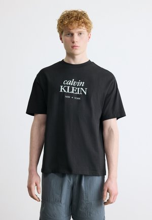 Tânăr cu păr roșcat și creț, purtând un tricou negru Calvin Klein și pantaloni scurți gri, stând pe un fundal alb simplu.