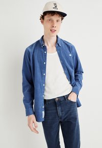 Μπλε πουκάμισο με κουμπιά από μαλακό ύφασμα, λευκή μπλούζα από μέσα, σκούρα τζιν και κρεμ καπέλο με γραφικό λαγού. Χαλαρή, άνετη εφαρμογή.