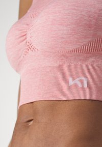 Sostén deportivo de color rosa claro con tela texturizada, banda inferior acanalada y detalle de malla. Presenta un pequeño logo blanco en la banda.