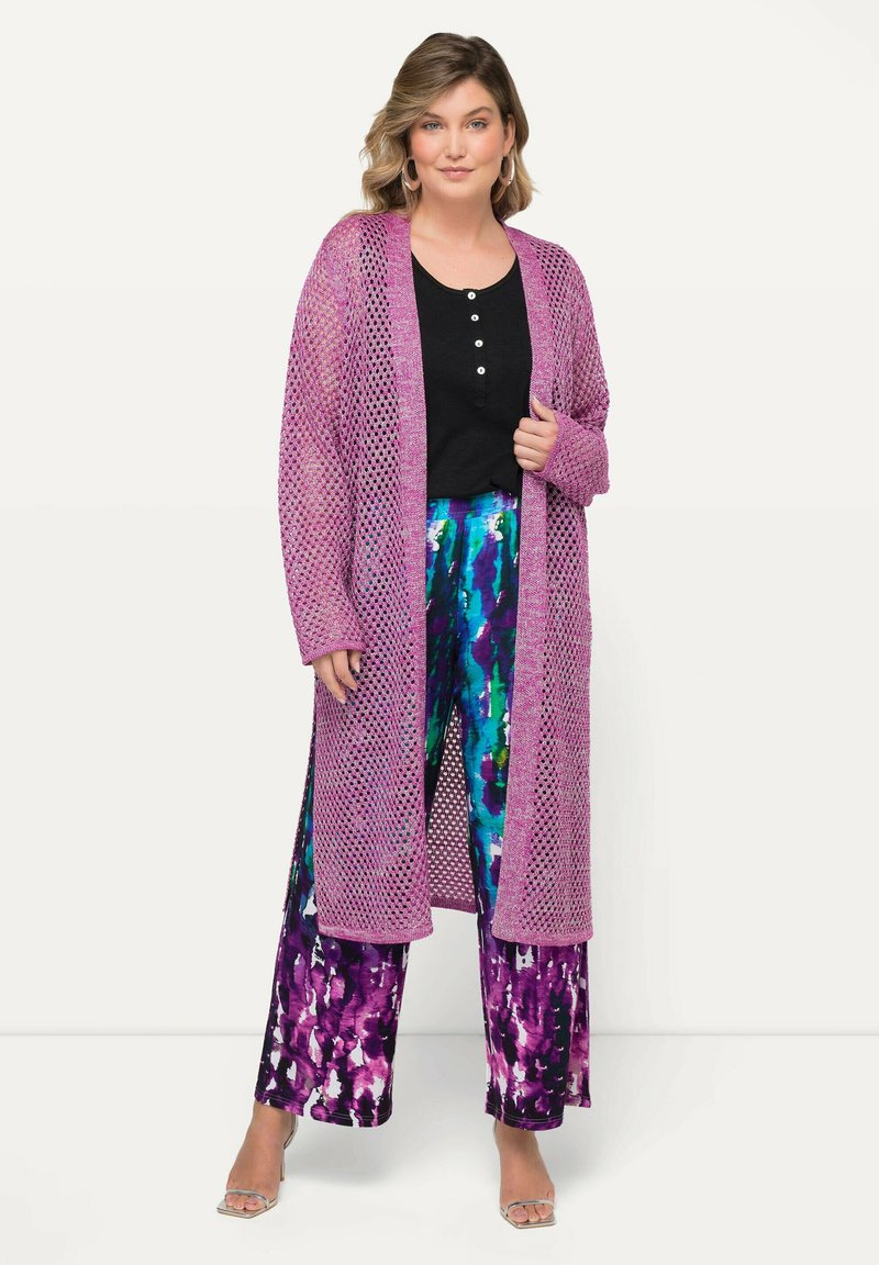 Cardigan largo de punto abierto en un vibrante color púrpura, superpuesto sobre una blusa negra de botones y pantalones de pierna ancha multicolores con un patrón de acuarela.