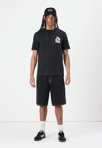 Sort kortærmet t-shirt med grafisk motiv på forsiden, parret med sorte denim shorts. Modellen bærer en sort kasket og sorte sneakers med hvide detaljer.