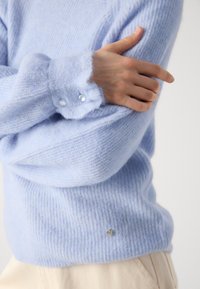 Lyseblå, ullblandingsgenser med stripete tekstur, detalj med knapper ved mansjettene og en liten metallisk aksent nær kanten. Myk og koselig utseende.