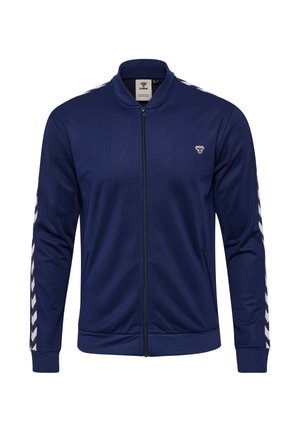 Mand og kvinde iført navyblå jakker med lynlås og v-formede striber på ærmerne; manden iført sorte shorts og sneakers, kvinden iført grå joggingbukser og hvide sneakers.