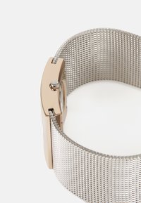 Montre dorée avec un cadran rond, bracelet en mailles argentées et un bouton sur le côté, présentant un design minimaliste et des textures lisses.