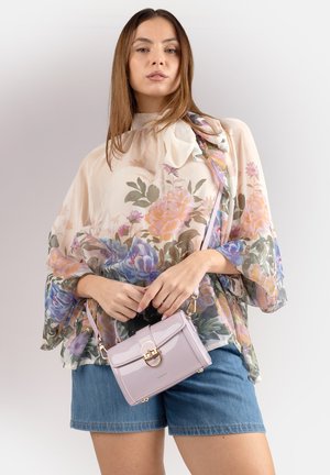 Donna con una blusa trasparente a fiori e pantaloncini di jeans, che tiene una piccola borsa rosa chiaro con una chiusura dorata.