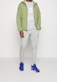 Chaqueta ligera verde, camiseta gris, pantalones de chándal grises y zapatillas deportivas azules. Detalles notables: bolsillos con cremallera y logos de marca.