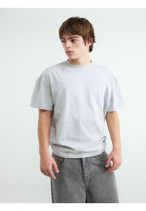 Mladý muž nosi svetlosivé voľné tričko a voľné sivé rifle, stojí pred jednoduchým bielym pozadím, pozerá doľava.