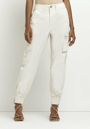 Femme portant un pantalon cargo blanc taille haute avec de grandes poches, des poignets élastiques, des fermetures éclair latérales et des sandales à talons hauts beiges à brides.