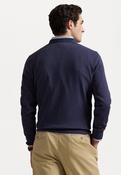 Pull marin avec une texture côtelée, doté d'un col et de manches longues, associé à un pantalon beige. Détails de conception subtils visibles à l'arrière.