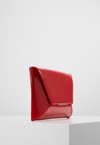 Pochette en cuir verni rouge brillant avec des panneaux latéraux triangulaires et un petit logo de marque doré rectangulaire sur le rabat avant.