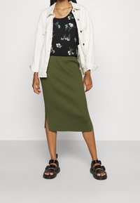 Grøn, ribbet midi-nederdel med en sideslids, parret med en sort blomstret tanktop og en hvid denimjakke. Sandaler med sorte stropper.