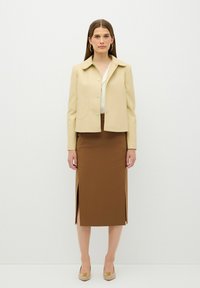 Beige Bikerjacke mit spitzem Kragen und grünen Knöpfen, kombiniert mit einem braunen, knielangen Rock mit seitlichen Schlitzen. Schuhe in neutralen Tönen.