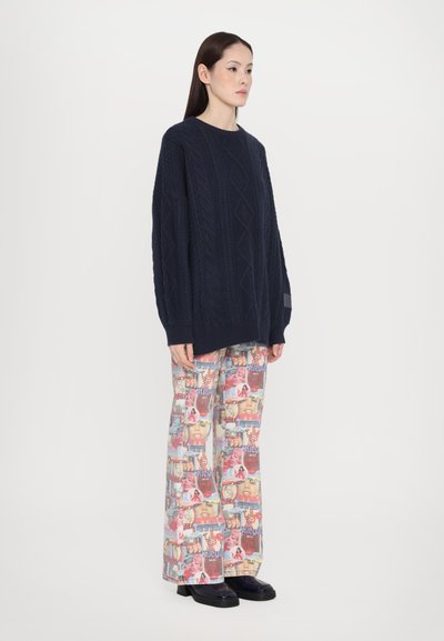 Pull en maille torsadée marine avec des motifs en losange, associé à un pantalon large à imprimés graphiques colorés. Des chaussures noires complètent la tenue.