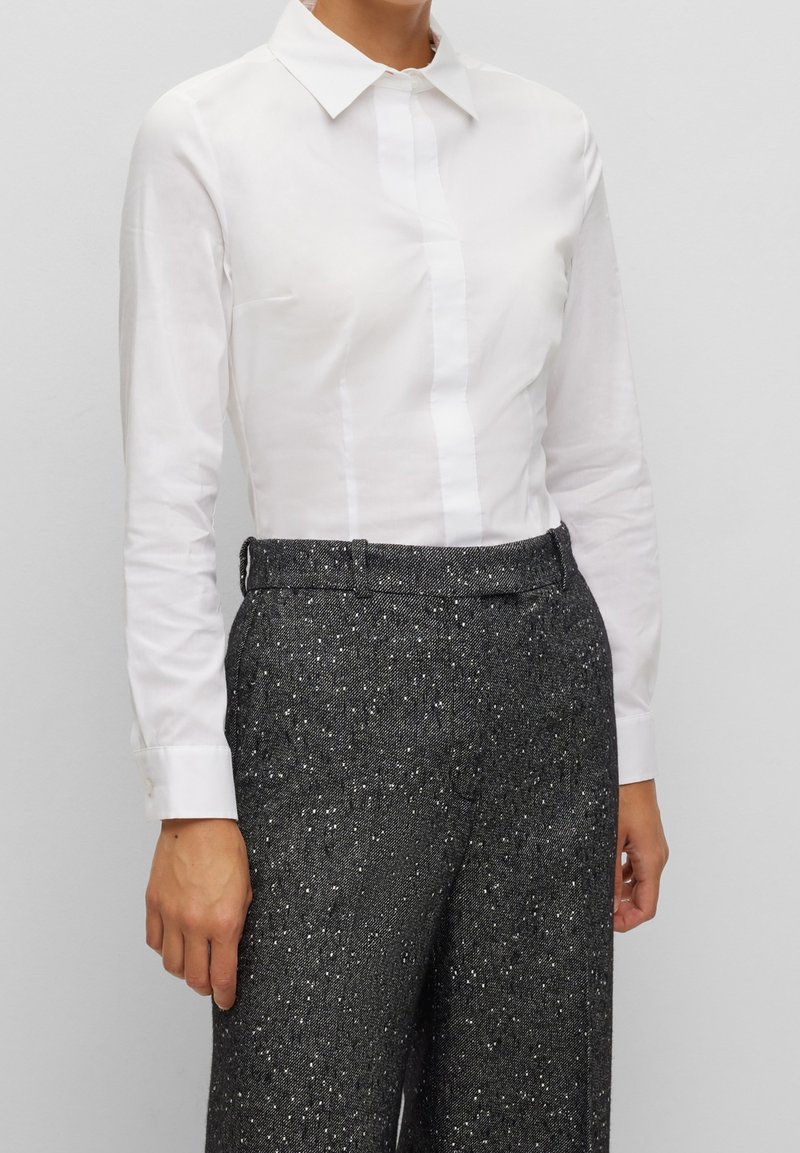 Camicia bianca a maniche lunghe con colletto classico e bottoni frontali, abbinata a pantaloni neri testurizzati con leggere sfumature argentate.