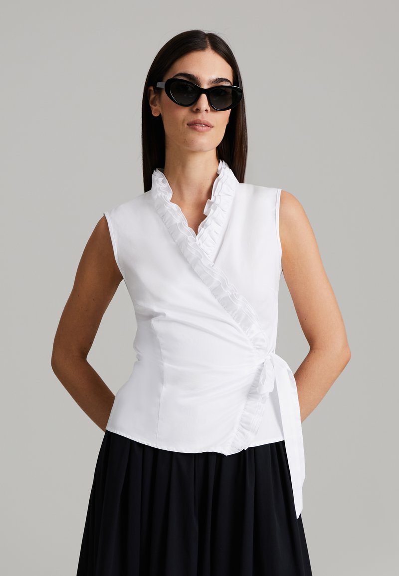 Top bianco senza maniche con colletto arricciato e dettaglio a fiocco in vita. Tessuto morbido, vestibilità sartoriale, abbinato a occhiali da sole neri.