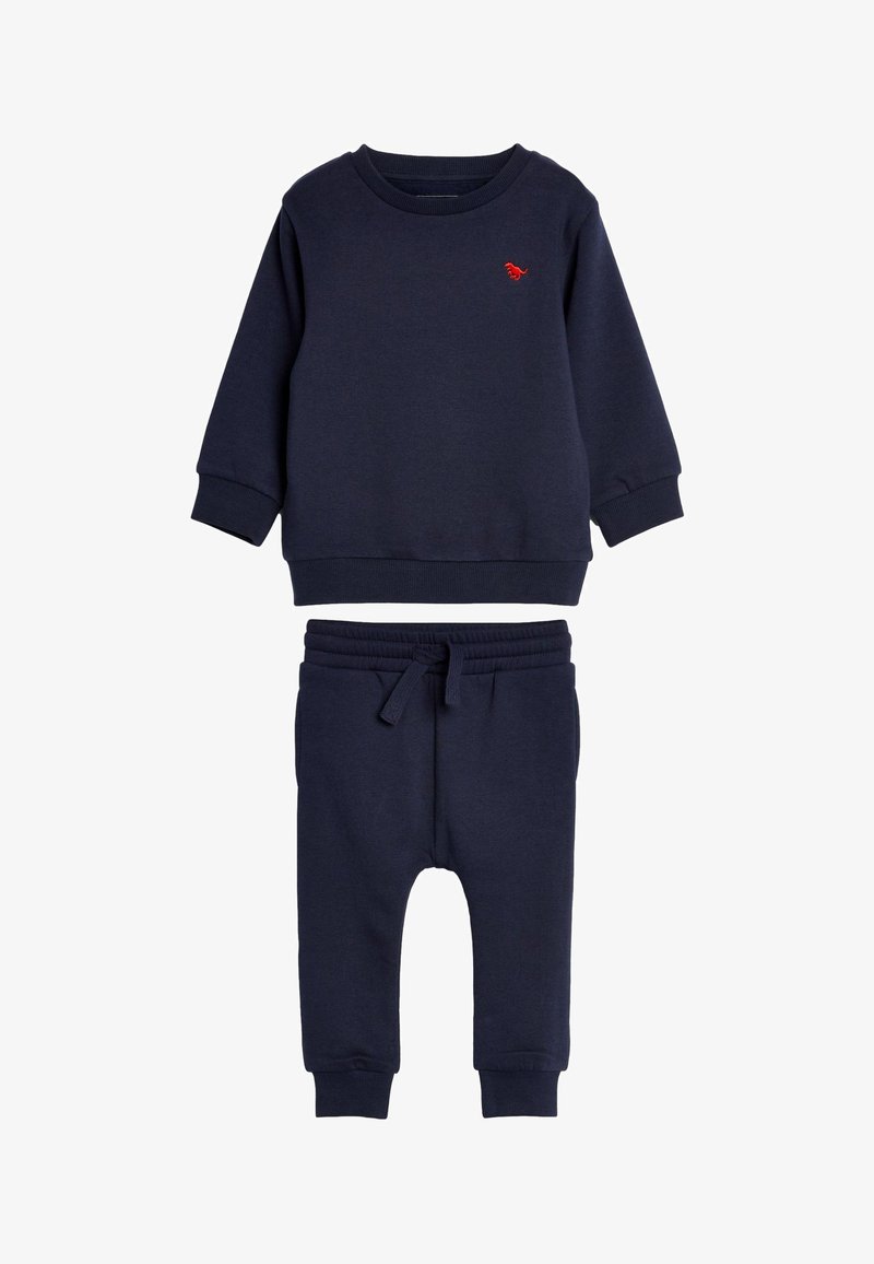 Tumši zila kokvilnas sporta krekla un bikses komplekts. Sporta krekls ir ar sarkanu izšuvumu; biksēm ir regulējama josta un ribotas aproces.