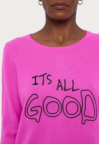 Femme portant un pull rose vif avec un texte brodé noir "ITS ALL GOOD" et des boucles d'oreilles créoles dorées, visible du cou au milieu du torse.