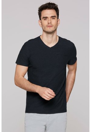 Young man with short dark hair wearing a black striped V-neck T-shirt and light grey pants, standing against a plain background.