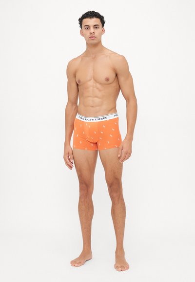 Muskulöser Mann steht barfuß in orangefarbenen Polo Ralph Lauren Boxershorts mit weißem Logo-Bund vor weißem Hintergrund.