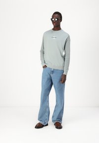 Maglione grigio a maglia con collo girocollo e dettaglio del logo, abbinato a jeans a gamba larga blu chiaro e scarpe in pelle marrone. Occhiali da sole neri indossati.