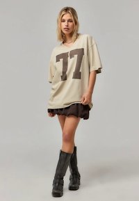 Overdimensioneret beige t-shirt med mørkebrun "77" tryk, parret med en rynket brun nederdel og sorte knæhøje støvler. Glat tekstur, afslappet design.