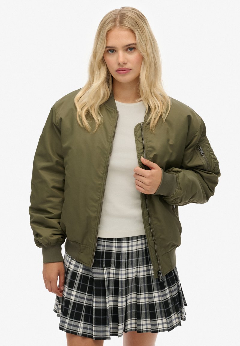Oliwkowa kurtka bomberka z zamkiem z przodu, ściągaczami przy mankietach i detalami kieszeni, zestawiona z plisowaną spódnicą w czarno-białą kratkę.