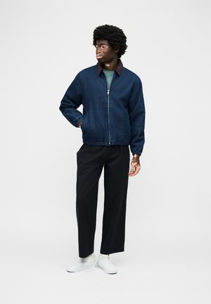 Νέος άνδρας με σγουρά μαλλιά, ντυμένος με σκούρο μπλε ζακέτα με φερμουάρ, μαύρο παντελόνι, λευκά αθλητικά παπούτσια, στέκεται με το ένα χέρι στην τσέπη κοιτάζοντας πλάγια.