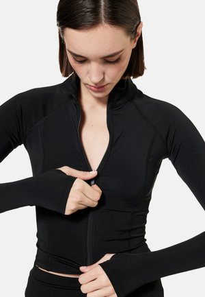 Nainen pukeutunut mustaan pitkähihainen vetoketjulliseen puseroon, jossa peukalonreiät, katsoo alas vetäessään vetoketjua ylös.