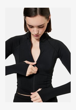 Nainen pukeutunut mustaan pitkähihainen vetoketjulliseen puseroon, jossa peukalonreiät, katsoo alas vetäessään vetoketjua ylös.