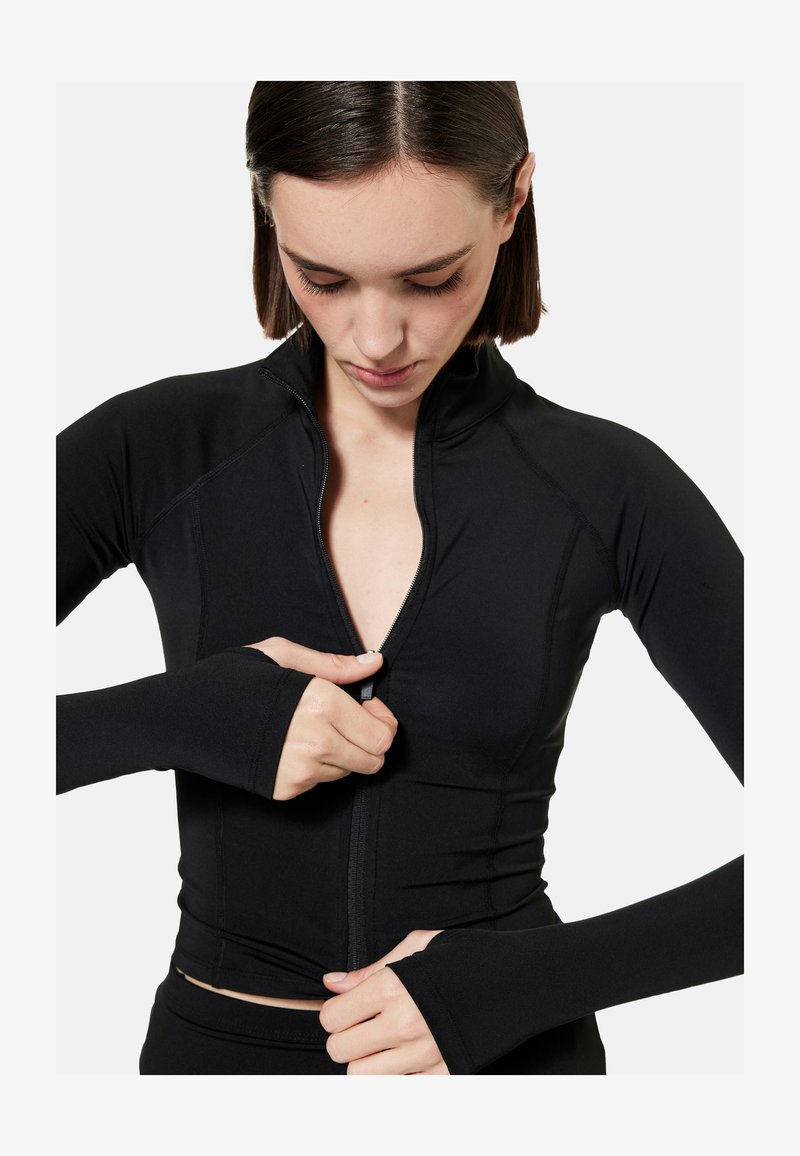 Woman wearing a black long-sleeve zip-up top with thumb holes, looking down while pulling the zipper upward.