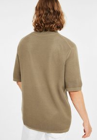 Persoon met schouderlang krullend haar die een gestructureerd olijfgroen kortemouwtrui en witte broek draagt, gezien vanachter.