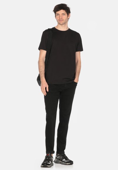 T-shirt nero a maniche corte, pantaloni neri slim-fit e scarpe sportive nere con dettagli grigi. Il modello sta in piedi dritto, tenendo una borsa.